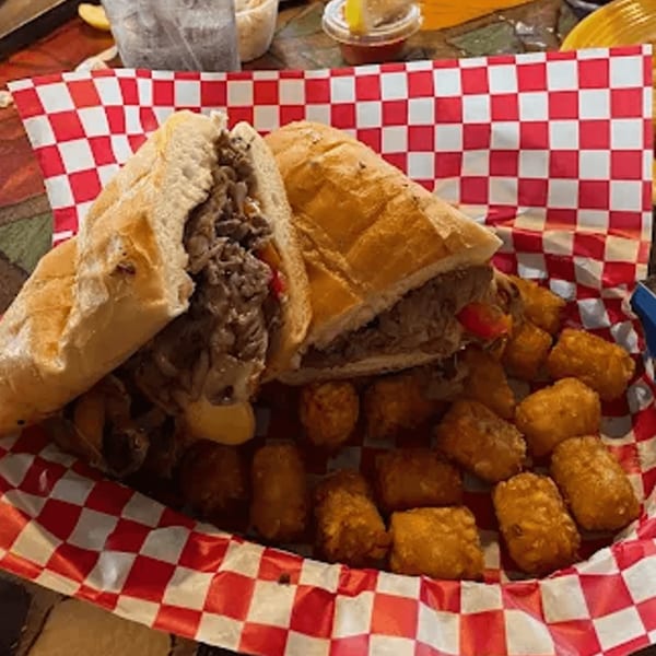 Cheesesteak sandwich with melted cheese and tater tots in a red and white checkered basket.