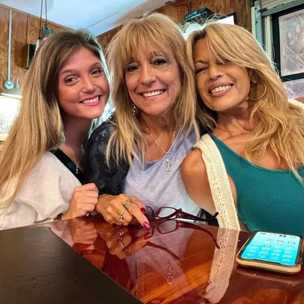 Three smiling women with blonde hair pose together at a wooden table in Graham's Landing.