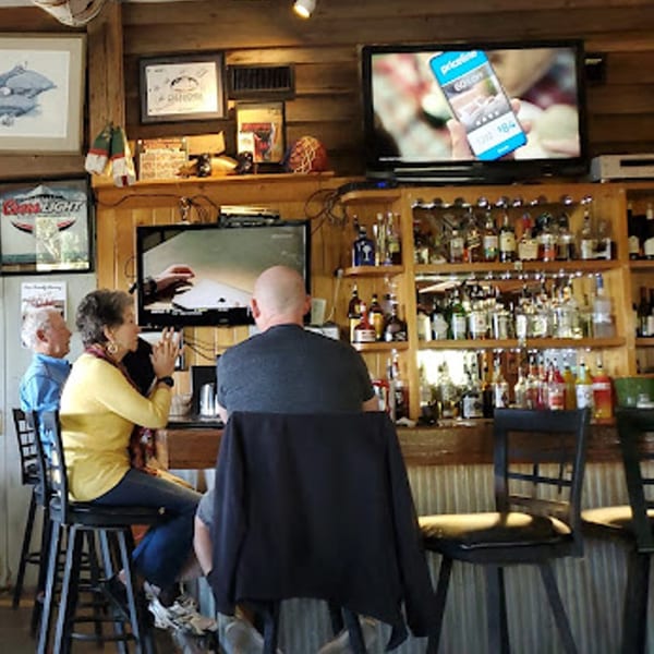 Patrons seated at a rustic wooden bar with shelves of liquor bottles and two TVs.