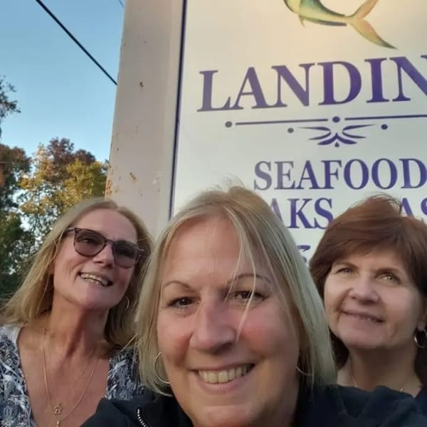 Three smiling women posing for a selfie in front of the Graham's Landing seafood restaurant sign.